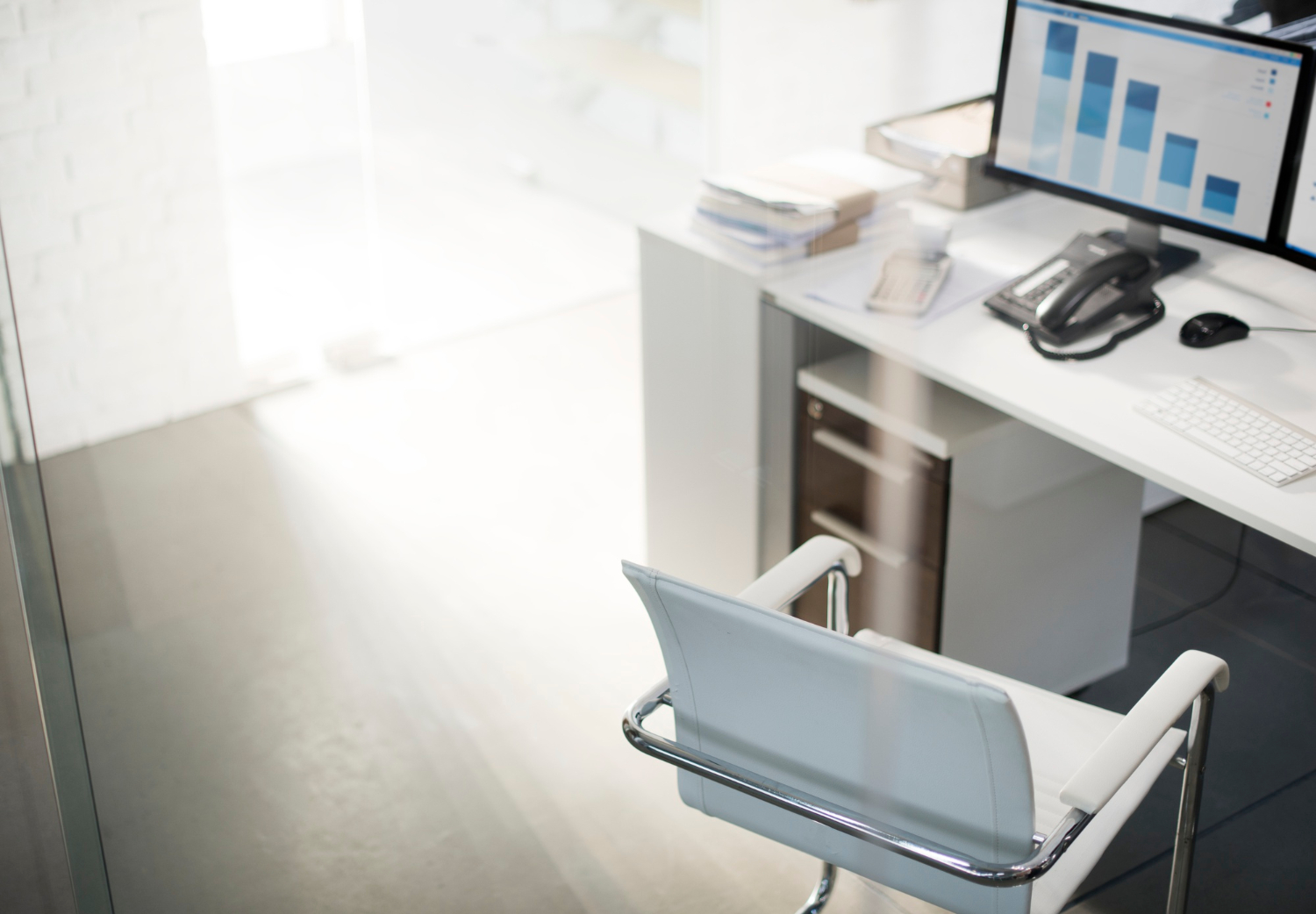 Escritório com mesa de trabalho, computador e cadeira branca, visto através de uma divisória de vidro